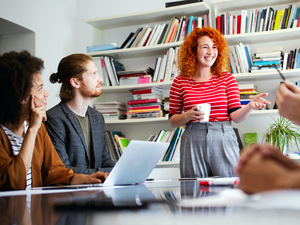 Photo of people having a business meeting, with one person standing up and presenting to the group. They are all wearing business casual clothing.