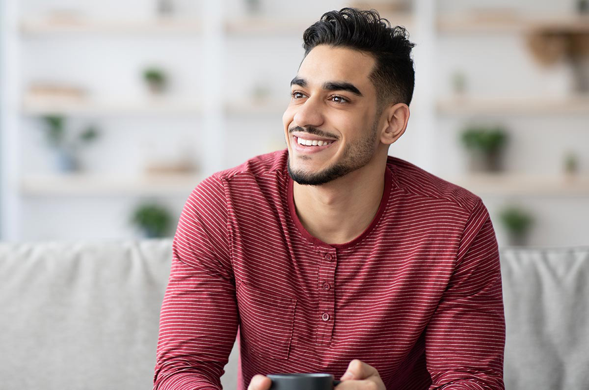 Photo of a person smiling. They're sitting on a couch in their living room, holding a cup of coffee.
